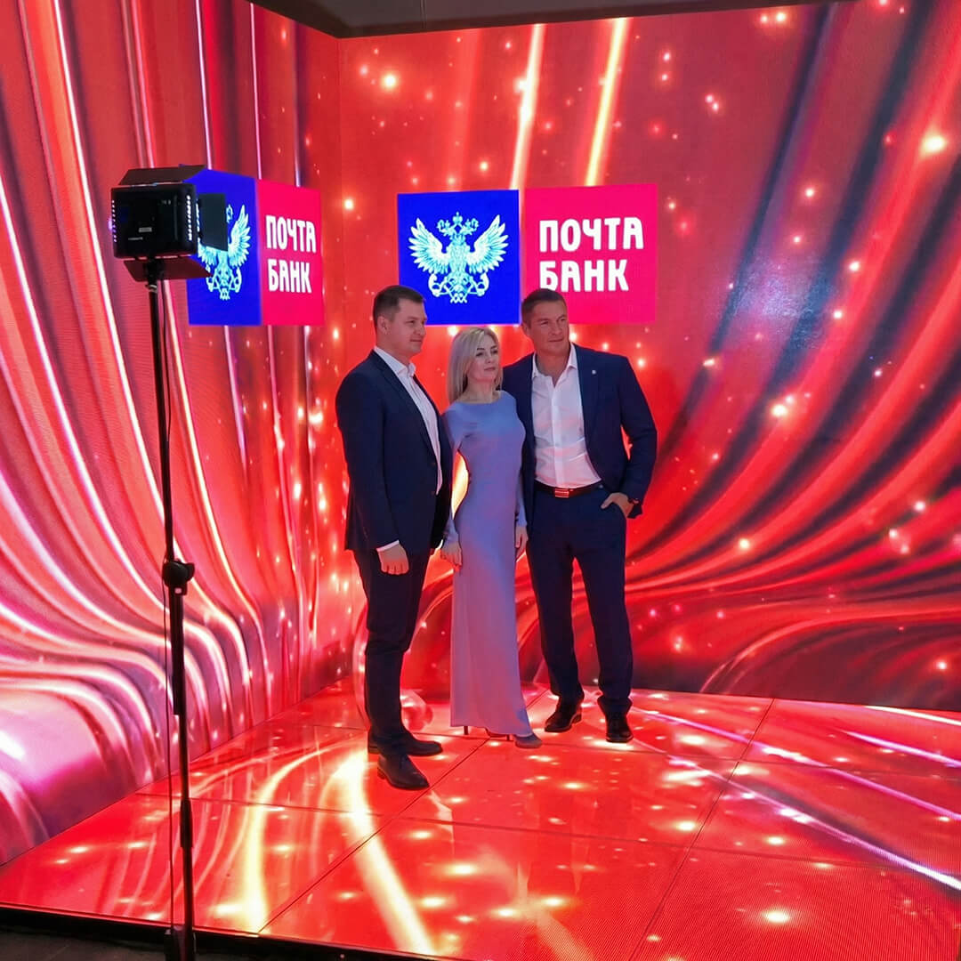 В угловой led фотозоне стоят два молодых человека в костюмах и девушка в платье на красном фоне и полу из экранов